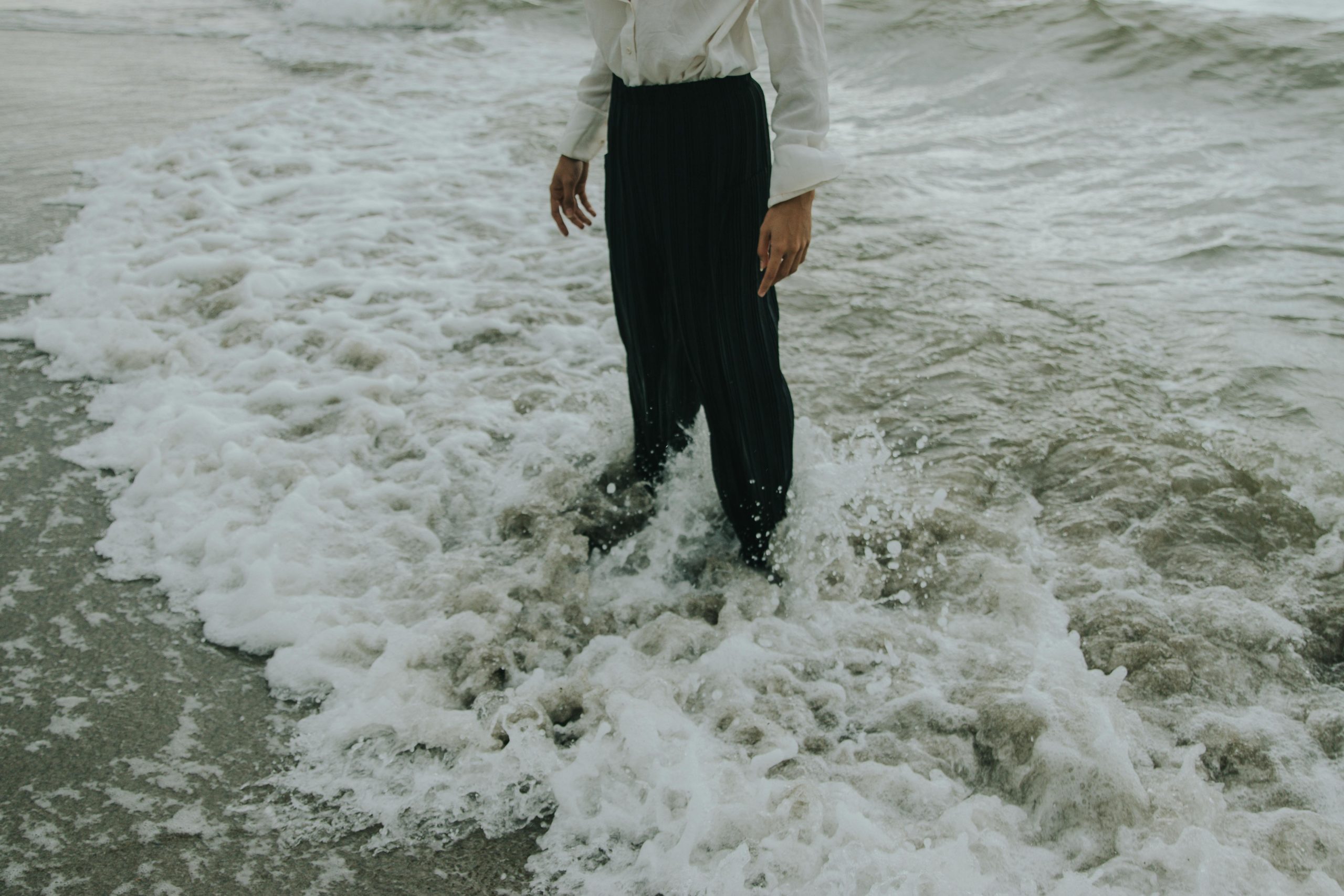 A person standing on a beach with waves splashing against their black pants, creating a serene coastal scene.